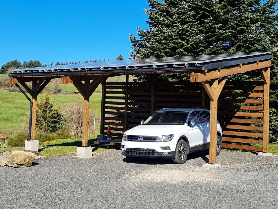 Carport en bois couvert de panneaux solaires abritant un SUV blanc, devant une prairie