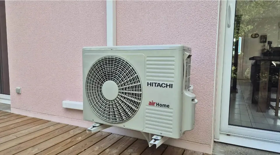 Groupe extérieur de climatisation Hitachi airHome fixé sur une façade rose au-dessus d’une terrasse en bois